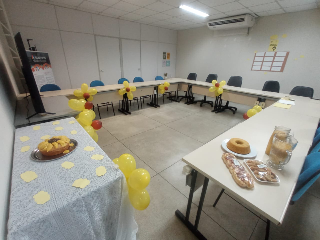 Esta é uma fotografia de uma sala de reunião decorada. À esquerda, há uma mesa coberta com uma toalha branca de renda, sobre a qual está um prato de sobremesa com fatias de bolo de cor amarela. No fundo da sala, há uma mesa em formato de 