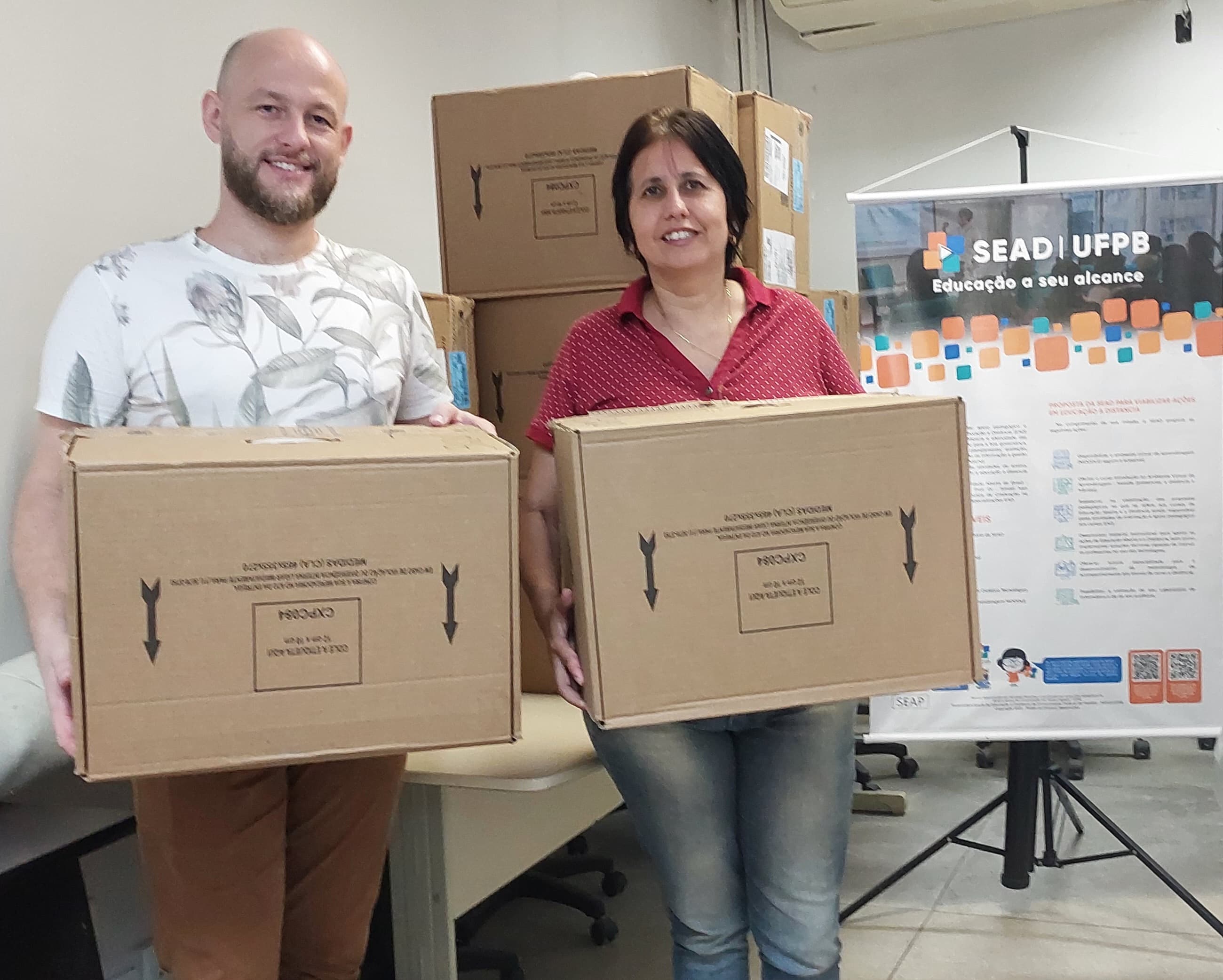 coordenadores da universidade aberta do brasil segurando caixas de papelão a esquerda, ao fundo uma pilha de muitas caixas de papelão, a direita um banner com informações sobre a superintendência de educação a distância - sead